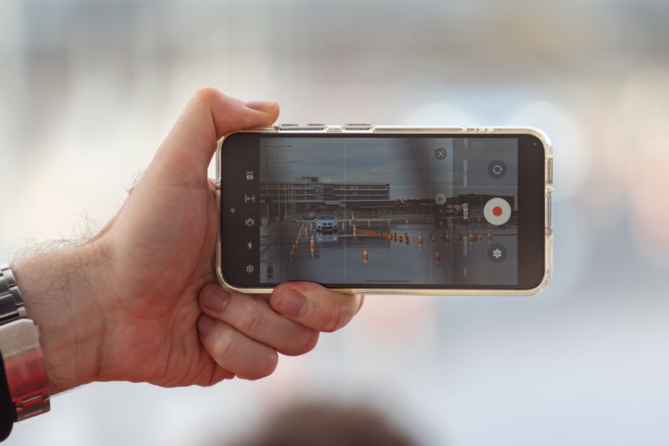 Jornalismo Cidadão — A hand holding a smartphone recording a video of a car scene outdoors. Captured in Balatonfőkajár
